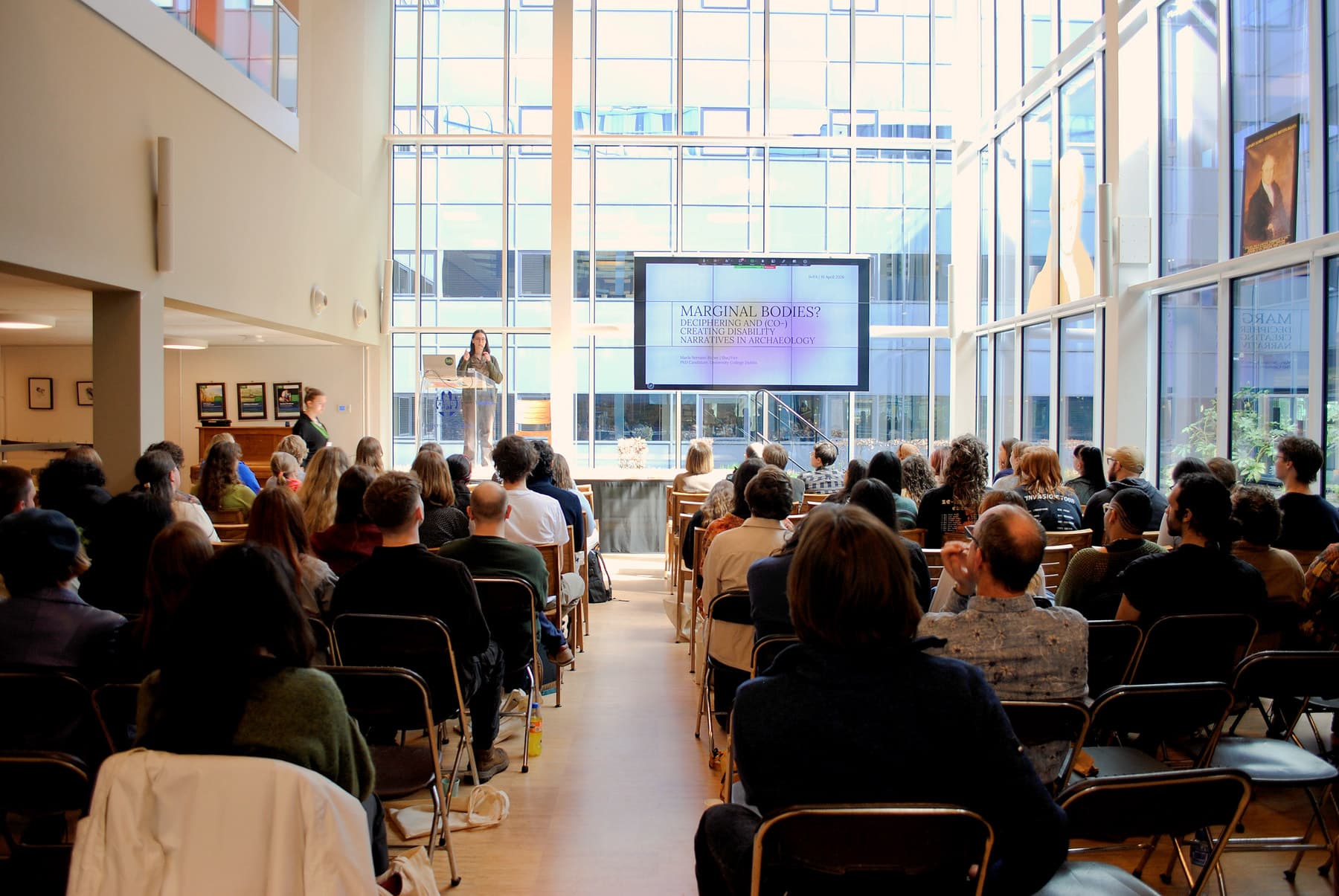 A speaker presents to a large seated audience in a sunlit glass atrium at SoYA 2026
